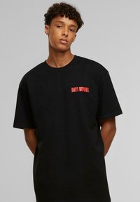 Sort bomulds T-shirt med korte ærmer, med teksten "DAYS BEFORE" i rød på venstre brystområde. Enkel rund hals design.