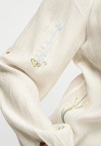 Μαλακό λευκό ύφασμα με διακριτικές κάθετες ρίγες που διαθέτει λεπτά κεντητά λουλούδια σε κίτρινο, μπλε και πράσινο.