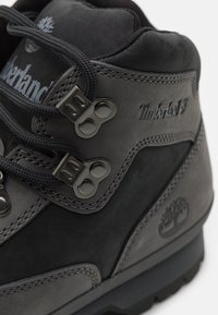 Juodi Timberland batai, pagaminti iš zomšos ir odos, su metaliniais akytėmis, tekstūruotais akcentais ir ryškiu logotipu šone.
