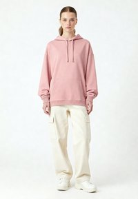 Felpa rosa oversize con tasca sul davanti e cappuccio con cordino, abbinata a pantaloni cargo bianchi con tasche laterali e sneakers bianche.