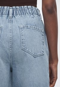 Ανοιχτό μπλε τζιν παντελόνι ψηλόμεσο με ελαστική μέση και πίσω τσέπες, φορεμένο από άτομο με σκουρόχρωμο δέρμα και καφέ τοπ.