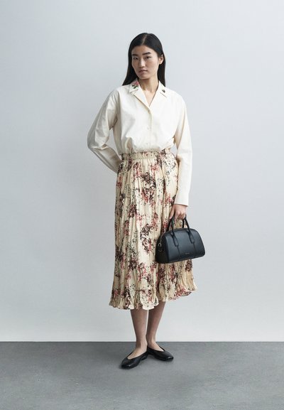 Mulher vestindo uma blusa creme com bordado floral na gola, uma saia midi plissada com estampa floral, sapatilhas pretas e segurando uma mala preta.