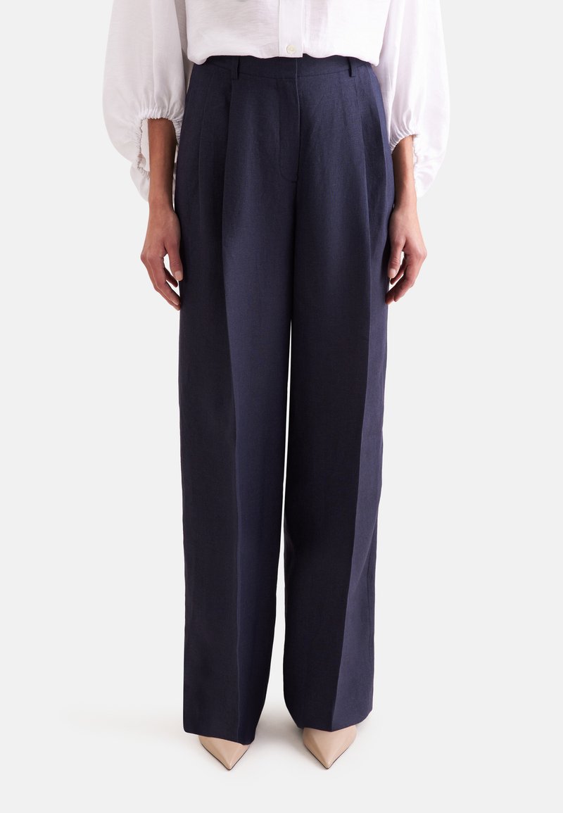 Femme portant un pantalon bleu marine taille haute à jambes larges, associé à des talons beige à bouts pointus et un chemisier blanc à manches longues.
