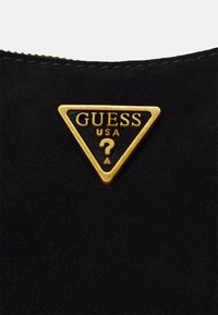 Czarna torebka zamszowa z trójkątną złotą blaszką z logo, na której znajduje się tekst "GUESS USA" oraz wzór znaku zapytania na środku.