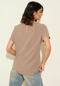 Top marrone a maniche corte con dettaglio a chiave sul retro, orlo arrotondato e texture liscia, abbinato a jeans baggy blu chiaro.