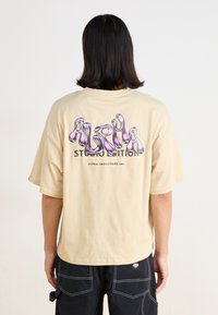 Beige oversized t-skjorte med en glanset lilla grafikk og teksten "ALPHA STUDIO EDITION" på baksiden. Korte ermer, laget av bomull.