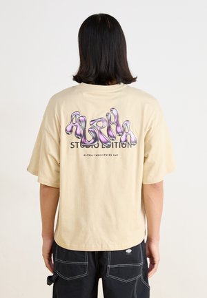 Beige oversized t-skjorte med en glanset lilla grafikk og teksten "ALPHA STUDIO EDITION" på baksiden. Korte ermer, laget av bomull.