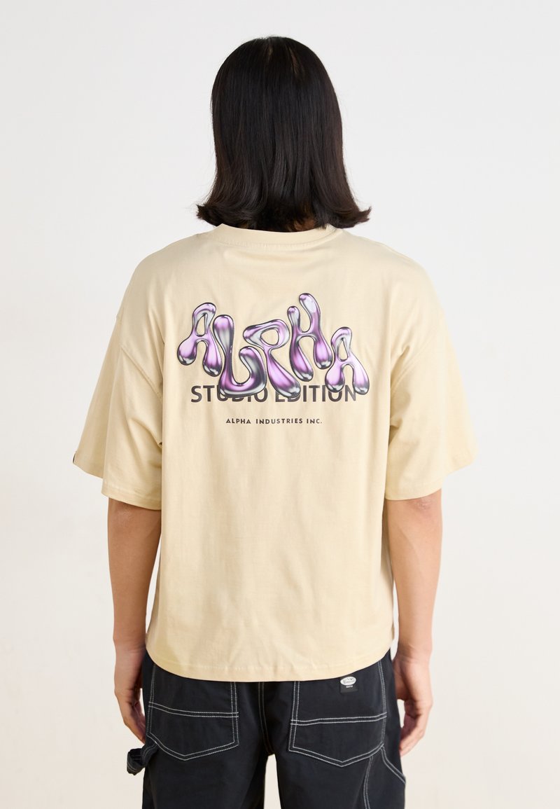 Beige oversized t-skjorte med en glanset lilla grafikk og teksten "ALPHA STUDIO EDITION" på baksiden. Korte ermer, laget av bomull.
