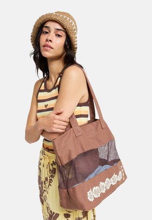 Kobieta ubrana w prążkowany top na ramiączkach, spódnicę we wzory i słomkowy kapelusz, niosąca na ramieniu brązową siatkową torbę z białym kwiatowym wzorem.