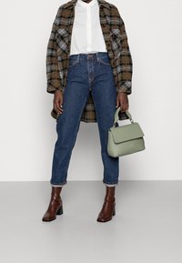 Personne portant un jean taille haute bleu, une chemise blanche à boutons, un manteau à carreaux, des bottes à talons brunes, tenant un sac à main vert sauge avec une poignée torsadée.