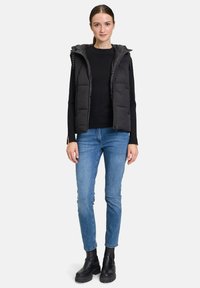 Schwarze gepolsterte Weste mit Kapuze über einem schwarzen Pullover, getragen mit hellblauen Jeans und schwarzen Stiefeletten. Verfügt über einen Reißverschluss und seitliche Taschen.