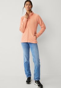 Fleece zip-up jack in zacht perzik met een hoge kraag, zijzakken en een getailleerd ontwerp. Gecombineerd met lichtblauwe jeans en zwarte sneakers.