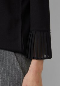 Schwarze Bluse aus strukturiertem Stoff mit plissierten, durchsichtigen Manschetten. Die Manschetten heben sich von dem glatten Hauptteil ab. Im Hintergrund graue Nadelstreifenhose.