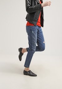 Giacca di pelle nera, camicia rossa, jeans blu strappati e scarpe nere testurizzate con dettaglio a fibbia, che mostrano un outfit casual in movimento.