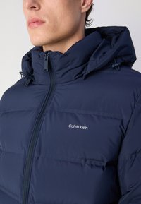 Tamno plava jakna s kapuljačom, punjena s materijalom koji drži toplinu, s prednjim patentnim zatvaračem i elastičnim podešavanjima, s logotipom "Calvin Klein" u bijeloj boji.