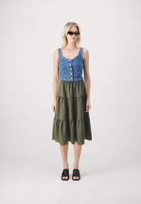 Cuerpo de denim con detalle de botones, falda acampanada en verde olivo y sandalias de dedo abiertas negras. Tejido suave y diseño casual.