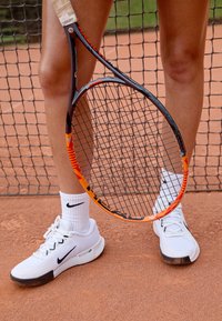 Hvite tennissko med sorte detaljer, hvite sokker og et tennisslag med en svart og oransje ramme, stående på en grusbane.