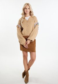 Cardigan beige in maglia con tre grandi bottoni e dettagli di pom-pom colorati sulle maniche, abbinato a una gonna di camoscio marrone e stivaletti alla caviglia color sabbia.