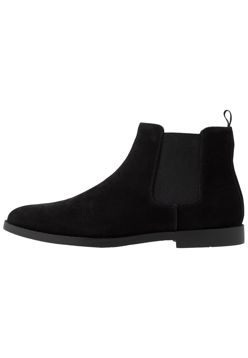 Stivale alla caviglia in suede nero con pannelli laterali elastici, una linguetta sul retro e suola in gomma piatta. Texture liscia e design minimalista.