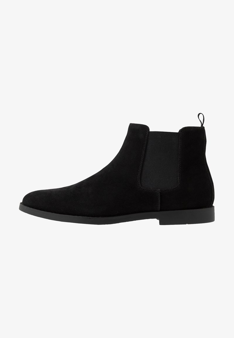Stivale alla caviglia in suede nero con pannelli laterali elastici, una linguetta sul retro e suola in gomma piatta. Texture liscia e design minimalista.