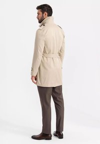 Beżowy płaszcz trench z wysokim kołnierzem, talią w pasie oraz guzikami na ramionach, w połączeniu z brązowymi spodniami i czarnymi botkami.