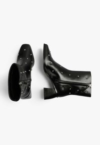 Botines de cuero negro con puntera puntiaguda, tachuelas plateadas, tacón grueso y cierre lateral para facilitar su uso. Superficie texturizada que realza el estilo.