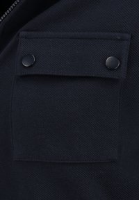 Giacca nera in tessuto con una tasca quadrata sul petto dotata di due bottoni a pressione neri e una superficie strutturata, che mette in mostra un design moderno e utilitaristico.