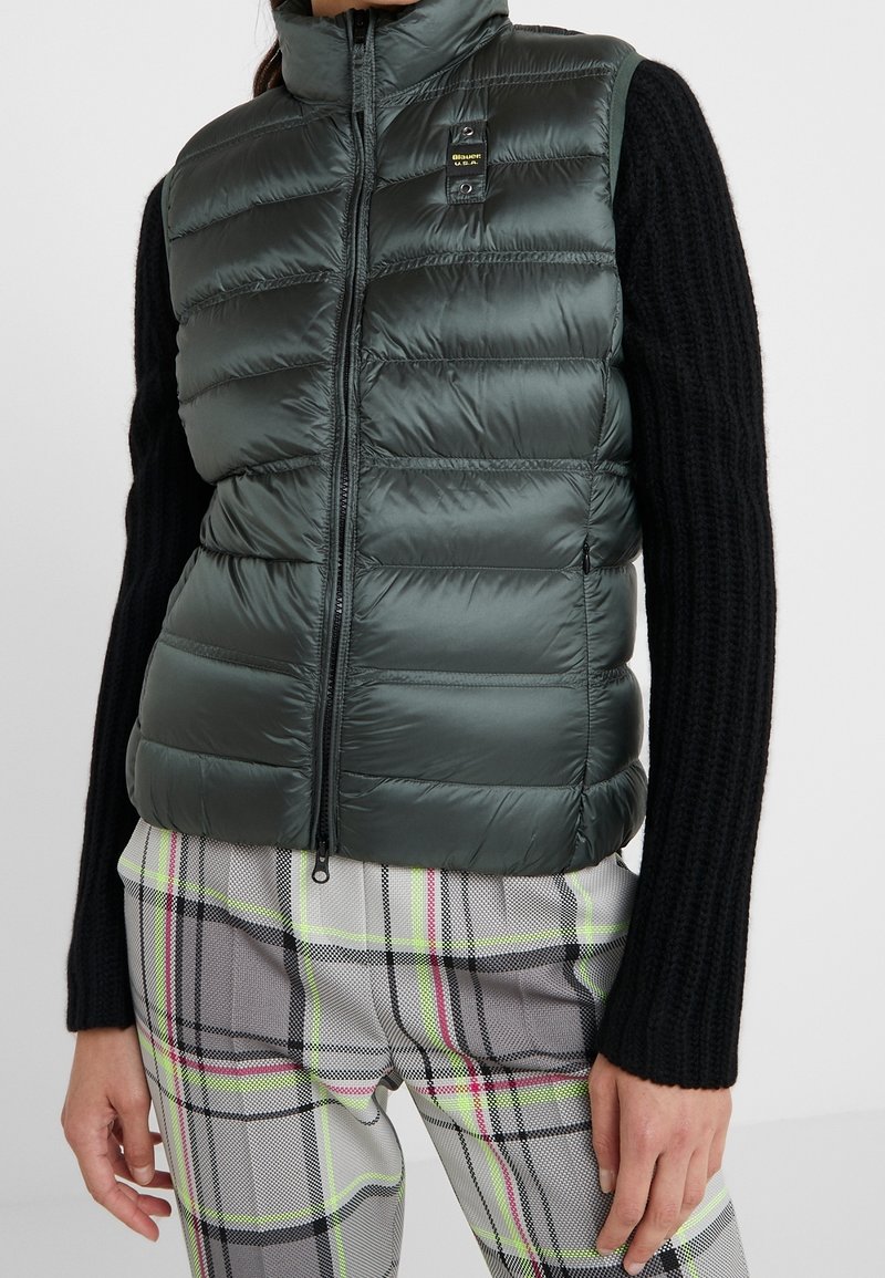 Grünes gepolstertes Westchen mit hohem Kragen, Reißverschluss und Seitentaschen. Getragen über einem schwarzen, gestrickten Langarmoberteil und karierten Hosen.