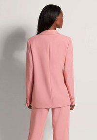 Lichtroze blazer met een getailleerde pasvorm, enkele split, gelapte revers en knoopaccenten op de mouwen, gecombineerd met bijpassende broeken.