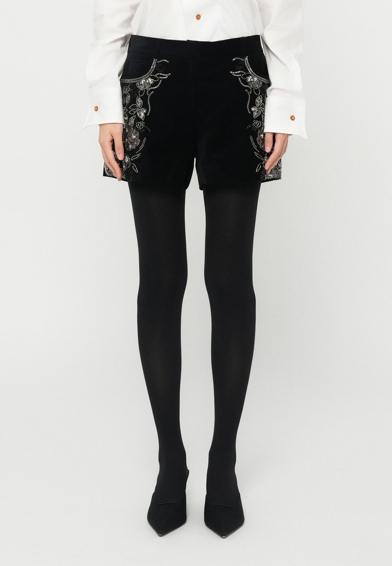 Fekete bársony sort szirommintás hímzéssel a zsebein, fekete harisnyával és hegyes fekete cipőkkel párosítva. Fehér, gombos blúz l�átható.