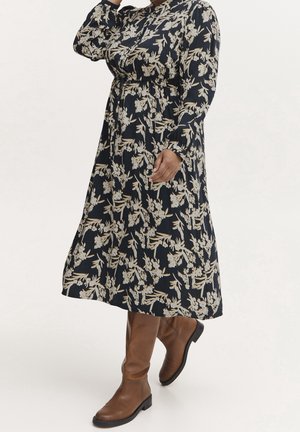 Kobieta w granatowej sukience midi w kwiaty z długimi rękawami i brązowych kozakach za kolano, pozująca z jedną ręką przy twarzy.