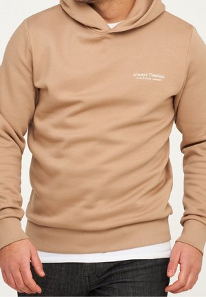 Sweat à capuche beige avec le texte « Always Timeless JACK & JONES x INFINITY » sur la poitrine, porté par-dessus une chemise blanche et un jean foncé.