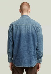 Denimskjorta i mellanblått. Har en krage med brett utfall, långa ärmar med knappmanschetter och en rak fåll. Bakifrån visas yoke-detaljen.