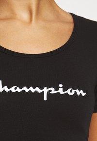 Černé bavlněné tričko s kulatým výstřihem a krátkými rukávy, s bílým kurzivním logem "Champion" tištěným na přední straně.