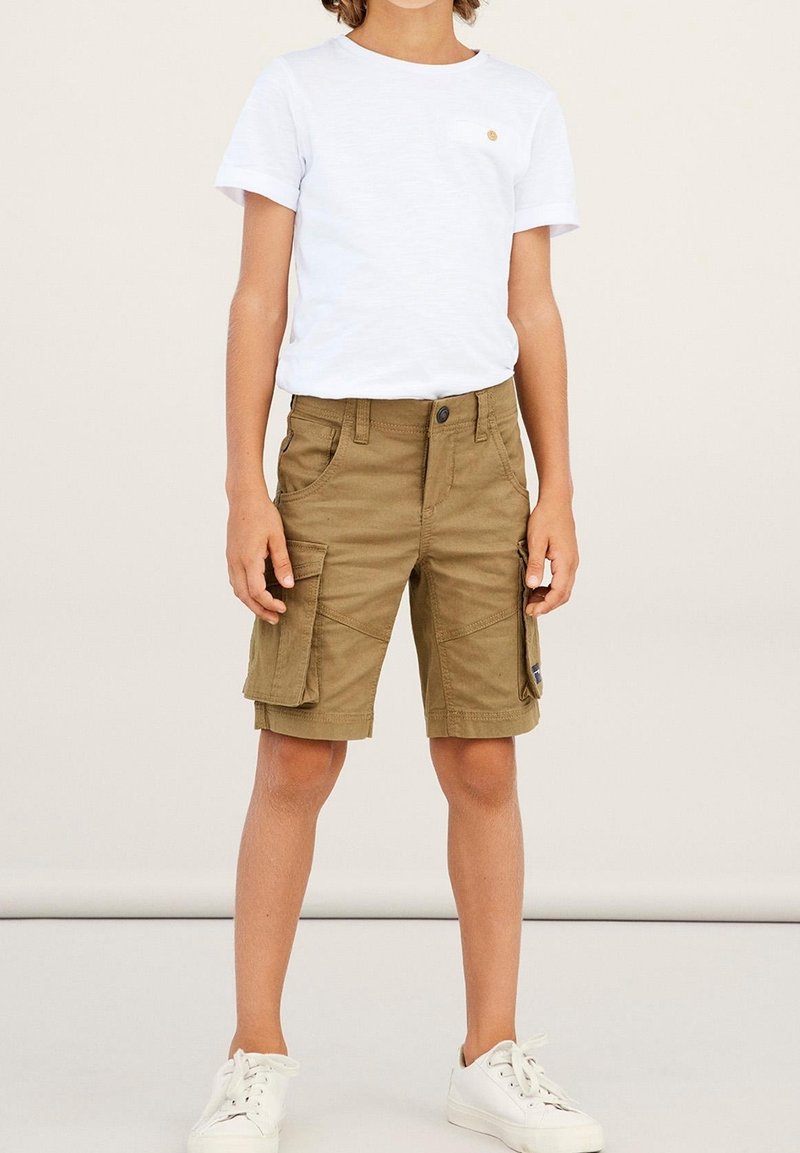 Enfant portant un t-shirt blanc à manches courtes, un short cargo marron avec des poches latérales, et des baskets blanches, debout devant un fond uni.
