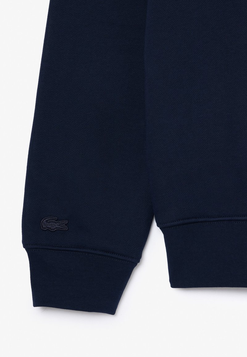 Granatowy sweter z ribbowanymi mankietami i dołem, posiadający teksturowany materiał oraz ciemny haftowany logo na lewym rękawie.