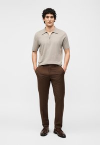 Νέος άνδρας που στέκεται με τα χέρια στις τσέπες, φορώντας μπεζ κοντομάνικο πόλο με φερμουάρ, καφέ παντελόνια και σκούρα καφέ επίσημα παπούτσια σε λευκό φόντο.