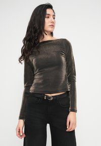 Μεταλλικό μπρονζέ τοπ με μακριά μανίκια και λαιμόκοψη καραβάκι, εφαρμοστό σχέδιο και απαλή υφή, συνδυασμένο με ψηλόμεσα μαύρα τζιν και μαύρη ζώνη.