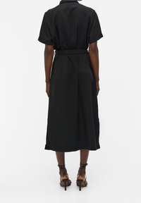 Schwarzes, kurzärmeliges Midikleid mit Gürtel, Rückenfalten und seitlichen Schlitzen. Glattes Material und lässige Passform, kombiniert mit hochhackigen Sandalen.