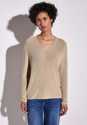 Pull léger beige en V avec texture côtelée et manches longues, associé à un jean en denim bleu. Style doux, décontracté et coupe ample.