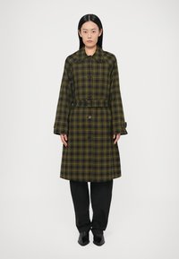 Pikk ruuduline mantel tumerohelises ja kollases toonis, krae, nööpidega kinnis, vööga vöörihm ja laiad varrukad. Kantud mustade pükste peal.