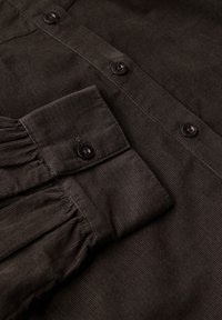 Il tessuto di velluto a coste marrone scuro presenta un design con bottoni, dotato di due bottoni e dettagli arricciati sulle maniche. La texture è a coste e liscia.