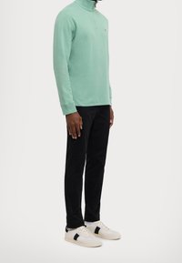 Man i mintgrön sweatshirt med kvartslång blixtlås, svarta byxor och vita sneakers med svarta ränder, som står framför en enkel vit bakgrund.