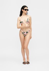 Femme portant un bikini rose à motifs tropicaux, des lunettes de soleil noires et des sandales noires à enfiler, debout avec un bras croisé sur son torse.