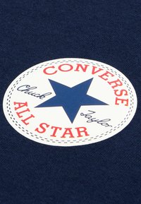 Sinine kangataust, millel on ümmargune valge plaastriga, millel on sinine täht, punasega "CONVERSE" ja mustas kursiivis "Chuck Taylor".