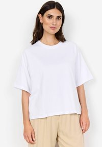 White cotton t-shirt with short sleeves and a crew neck, featuring a relaxed fit and smooth texture. Model wears beige trousers.