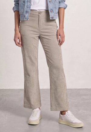 Femme portant un pantalon large en velours côtelé beige, des baskets blanches, un haut blanc et une veste en jean bleu avec les manches retroussées.