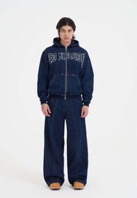 Denim-Zip-up-Hoodie mit großen weißen Texten, marineblaue weite Jeans mit sichtbaren Nahtdetails und beige Slipper.