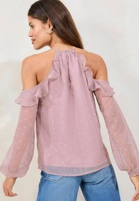 Puuvilla-lilac-payetilla kullanen pistekuvio, röyhelöillä varustettu avokainaladesign ja pitkät puffihihat, yhdistettynä vaaleansinisiin farkkuihin.