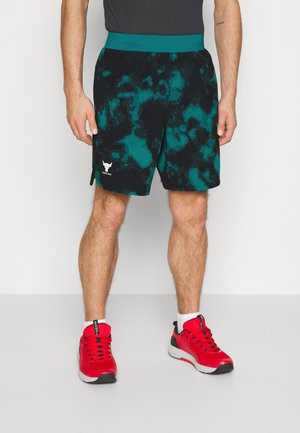 Mann trägt schwarze und türkise Camouflage-Sportshorts und leuchtend rote Laufschuhe mit weißen Socken, steht vor einem einfarbigen Hintergrund.